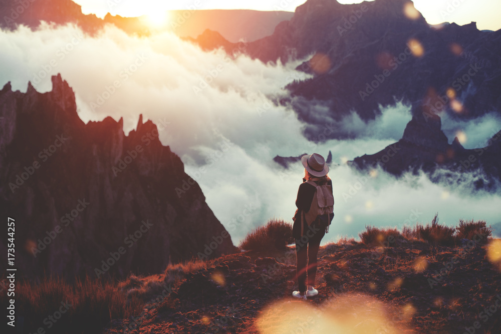 Young curly woman wearing  backpack, sneakers and hat standing on edge of cliff and enjoying landscape, sunset, mountains and clouds.