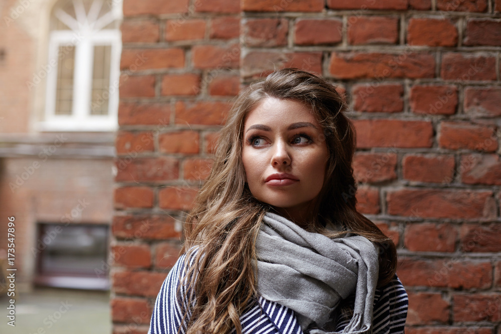 Style, fashion, people and modern urban lifestyle concept. Picture of fashionable young Caucasian female model wearing grey trendy scarf, standing at brick wall and looking away with mysterious smile Style, fashion, people and modern urban lifestyle concept. Picture of fashionable young Caucasian female model wearing grey trendy scarf, standing at brick wall and looking away with mysterious smile