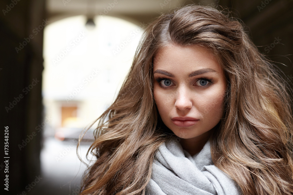 Close up lifestyle portrait of attractive young European female with voluminous hair and natural make up posing outdoors against arch background, looking at camera with serious facial expression Close up lifestyle portrait of attractive young European female with voluminous hair and natural make up posing outdoors against arch background, looking at camera with serious facial expression
