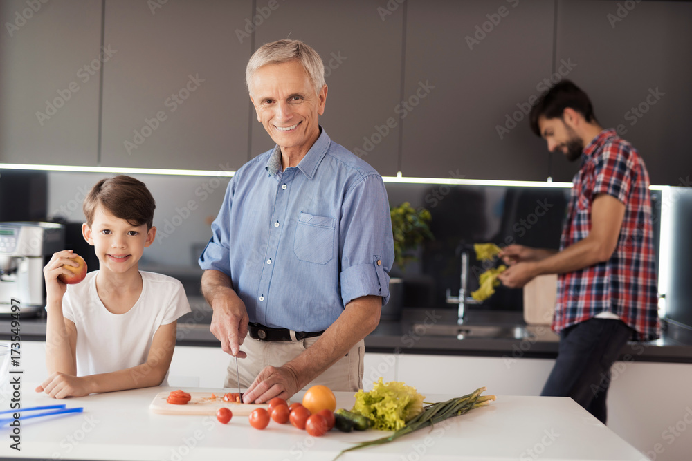 The old man and the boy are posing in the kitchen for making salad. The man behind them washes the lettuce leaves
