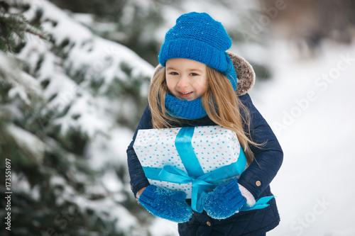 Little girl holding a christmas gift in her hands and emothionally smiling. Child wearing a blue knitted hat, gloves, mittens, scarf photo