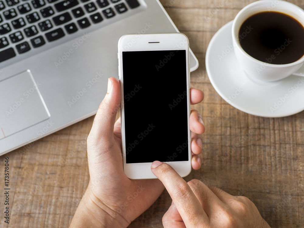 Close up business man's hand holds smart phone with black isolated screen over background of notebook, coffee on wooden background