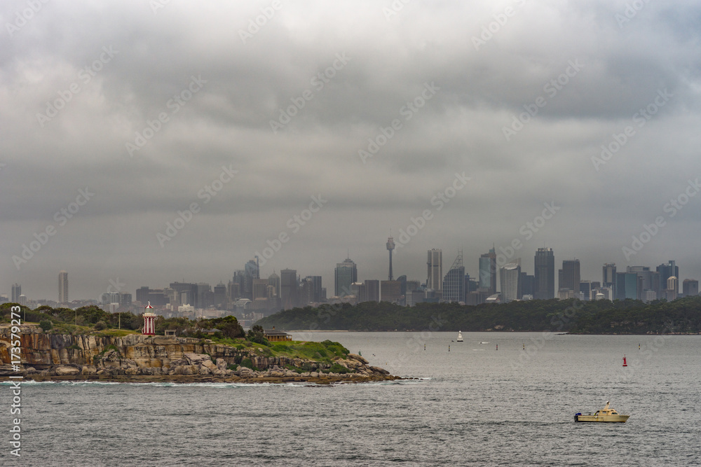 Sydney, Australia - March 21, 2017: South Head cliffs and park with short Hornby lighthouse up front, backed by Sydney skyline under foggy covered cloudscape. Small boat and buoys.