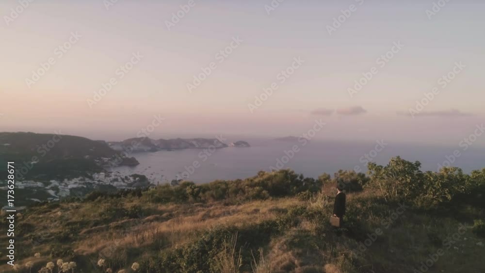 Business man in elegant suit walks on top of a mountain at sunset with the sea on the background
