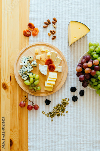 Top view pieses of cheese, grapes and dried fruits, nuts, pumpkin seedson wooden cheese plate on wooden table with strip tablecloth. Lay flat