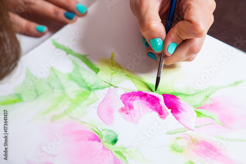 Close-up of the artist draws watercolor in the album for drawing with a thin wooden brush, on the table a pallet for drawing and a mug with tools