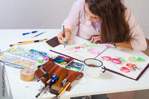 Dark-haired artist with bright draws in an album for drawing a thin wooden brush with pink watercolors, on the table lies a leather case with brushes, a mug of water, a palette of watercolors