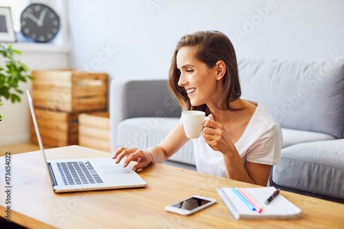Young woman drinking coffee and using laptop while sitting behind table at home