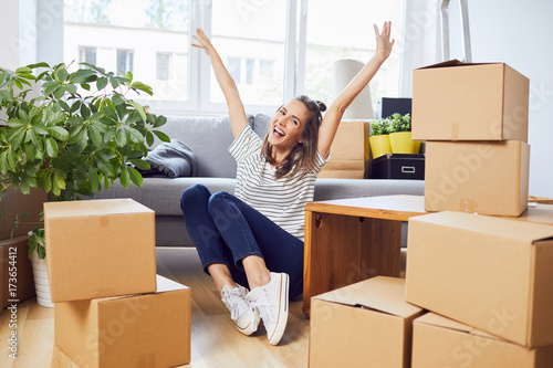 Ecstatic young woman sitting on floor in new apartment with boxes and raising arms in joy