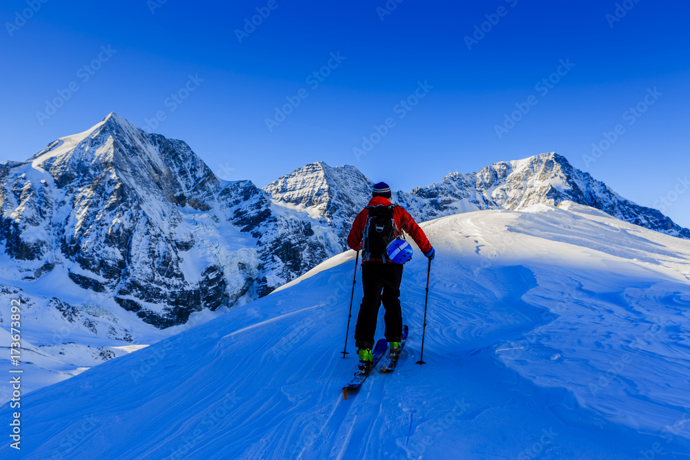 Mountain ski walking up along a snowy ridge with skis in the backpack. In background blue cloudy sky and shiny sun and Tre Cime, Drei Zinnen in South Tirol, Italy. Adventure winter extreme sport.