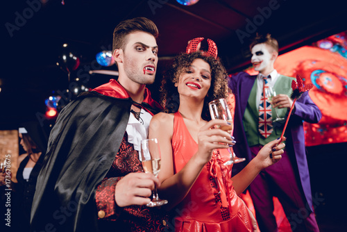 A guy dressed as a vampire and a girl dressed as a demon posing with champagne glasses in their hands and have fun