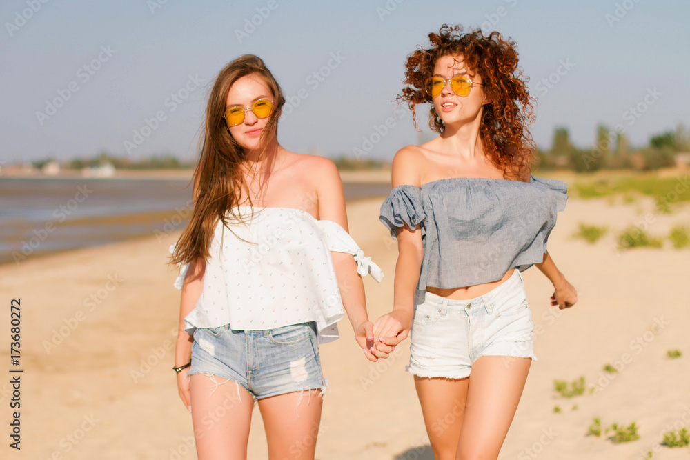 Portrait of two young female friends walking on the sea shore looking at camera laughing. Multiracial young women strolling along a beach. Portrait of two young female friends walking on the sea shore looking at camera laughing. Multiracial young women strolling along a beach.