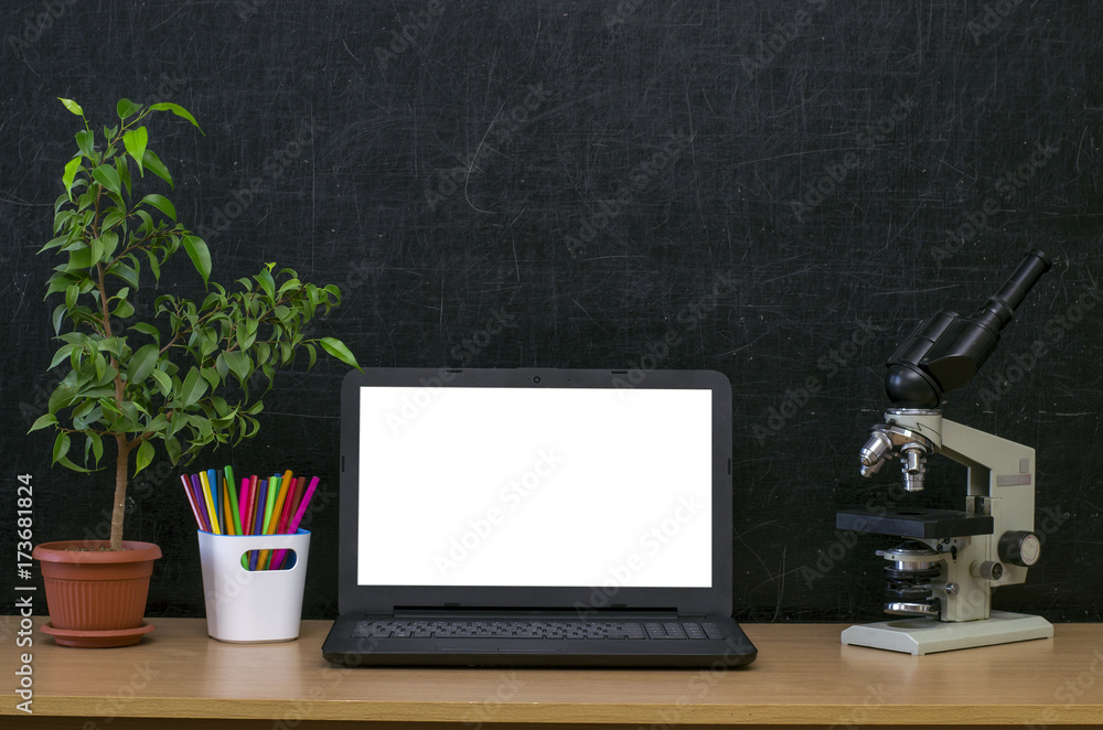 Fototapeta premium Teacher or student desk table. Education background. Education mockup concept. Laptop with blank screen, microscope, plant tree in the pot and colour pencils on blackboard (chalkboard) background.