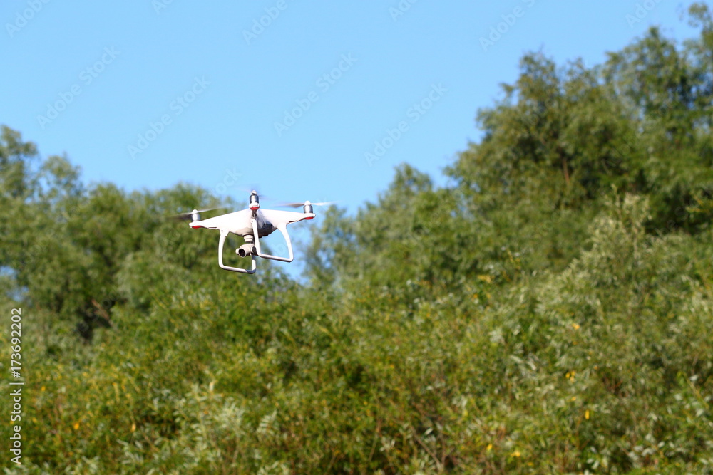 Drone with the camera against the blue sky and green trees. The flight of the copter in the sky.