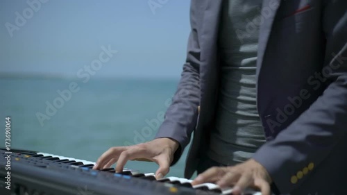 Hands, fingers of musician on digital piano. Young man performs witm synthesizer on azure coast, beach or quay. Male musician keyboardist plays music, song, have fun close to sea or ocean in summer photo