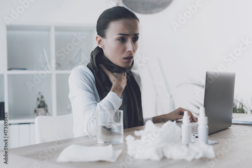 Sick woman sitting at her workplace in the office. She has a sore throat and she wore a scarf