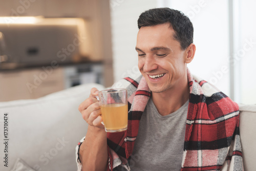 A man with a cold sits on the couch, hiding behind a red rug. He drinks medicinal tea