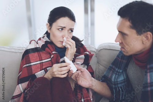 The couple is sitting on the couch wrapped in blankets. Man and woman are sick. A man gives a woman a nasal spray