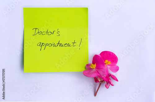 Doctor's appointment handwritten as a reminder on a yellow post it paper and a pink flower as decoration on white background