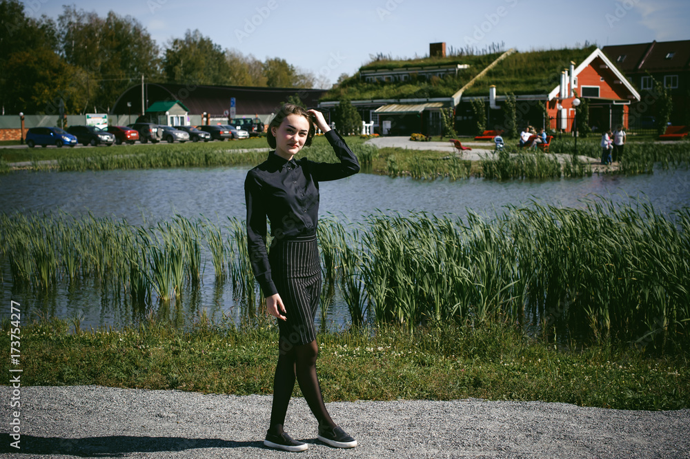 street portrait of a student girl dressed fashionably in dark clothes, against a pond background in the autumn sunny day