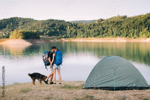 Hiking couple. Young couple with backpacks and dog kissing beside lake 