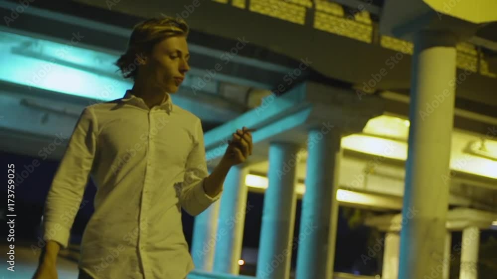 at night young blond-haired man walking along highway under bridge taking cigarette over his ear slow motion at night young blond-haired man walking along highway under bridge taking cigarette over his ear slow motion