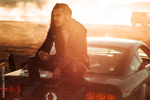 A man sits smoking on the back of his car in the desert sunlight photo