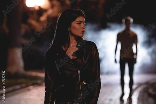 The woman stand on a dark street on a background of the man in fume. night time