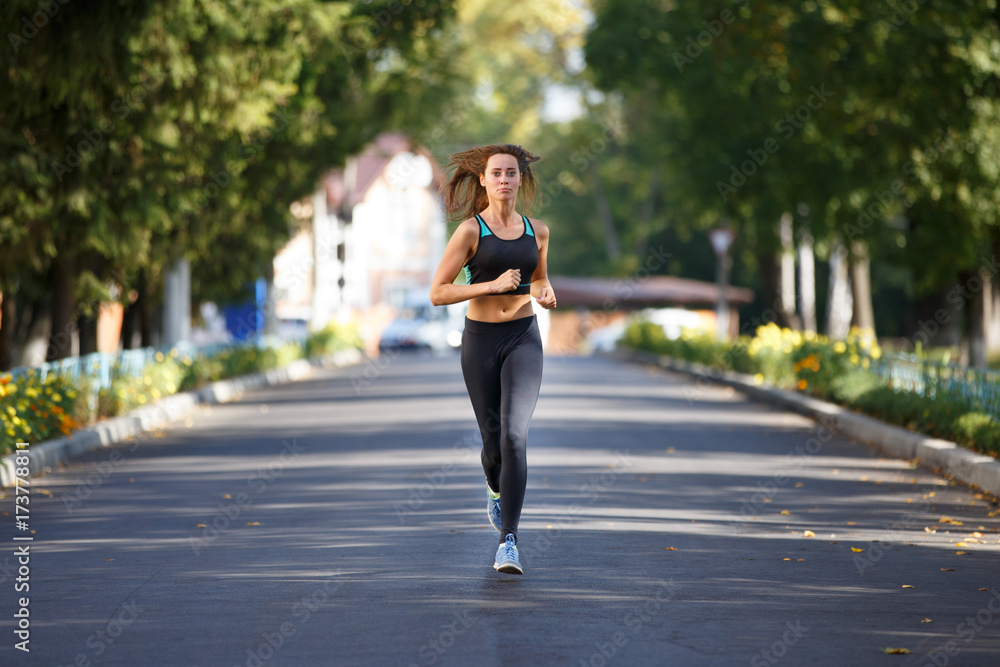 Young smiling sporty woman running on the road in the morning. Fitness girl jogging in park