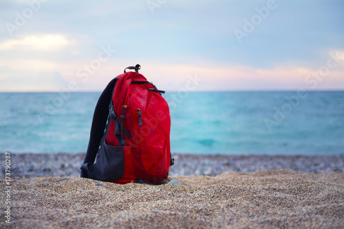 Red backpack for traveling stands on a sandy sea shore on the background of blurred sea. Horizontal Sea landscape