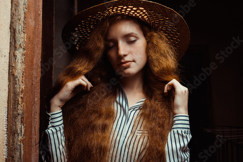 Tender redhead model with closed eyes in straw hat posing in passage. Closeup shot of redhead woman with long hair