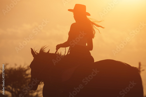 Leinwand Poster Sunset silhouette of young cowgirl riding her horse