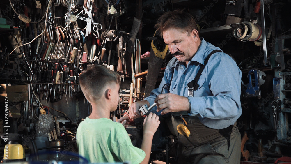 Close view of grandpa carpenter shows his grandson different tools in a small workshop. Close view of grandpa carpenter shows his grandson different tools in a small workshop.