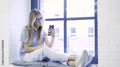 Blonde woman with long hair wearing glasses is sitting on a window sill and making a selfie. Handheld real time establishing shot photo