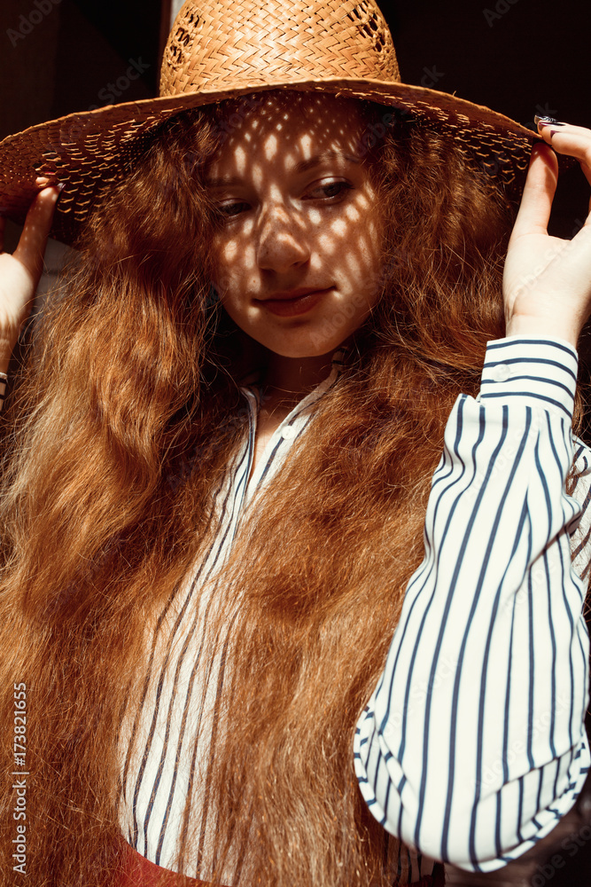 Fototapeta premium Elegant young red haired model in straw hat with shadow on her face