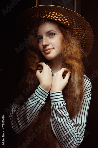 Lovely redhead model with long curly hair in straw hat posing near the old door