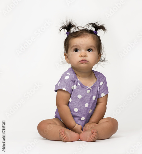 Sweet, serious baby girl sitting in polka dot onesie on white background