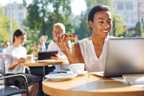 Cheerful woman holding croissant and reading from laptop
