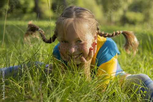 smiling blond girl with funny pigtail sits in grass. She is wearing a yellow T-shirt and a blue sarafan, and she has freckles on her nose.