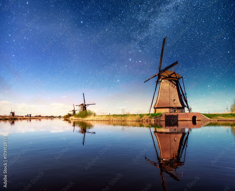 Colorful spring night with traditional Dutch windmills canal in Rotterdam. Wooden pier near the lake shore. Holland. Netherlands. Fantastic starry sky and the milky way.