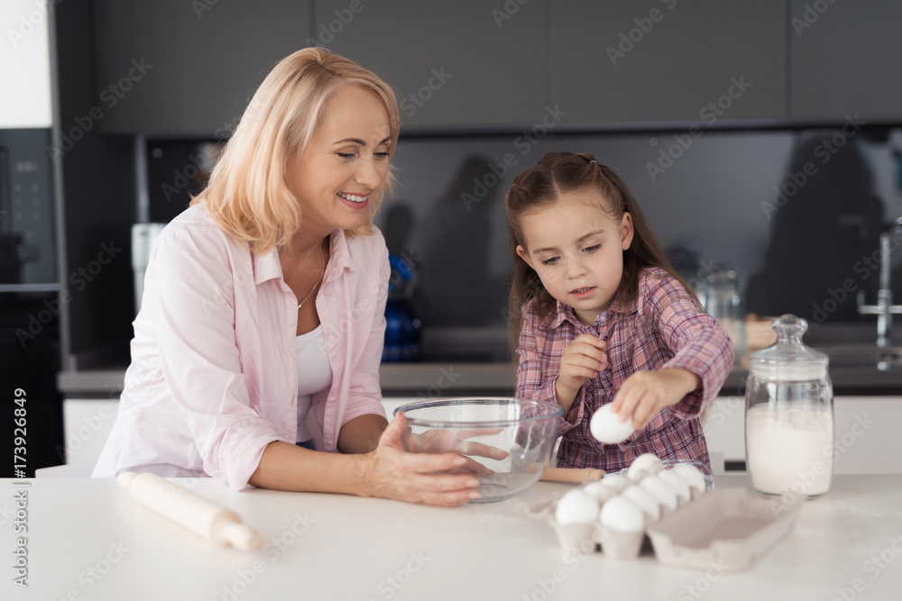 The girl and her grandmother are cooking pastries. The girl takes the eggs from the tray, the grandmother helps her