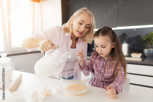 A little girl is cooking a homemade biscuit. The girl has beaten eggs, the grandmother pours milk in eggs