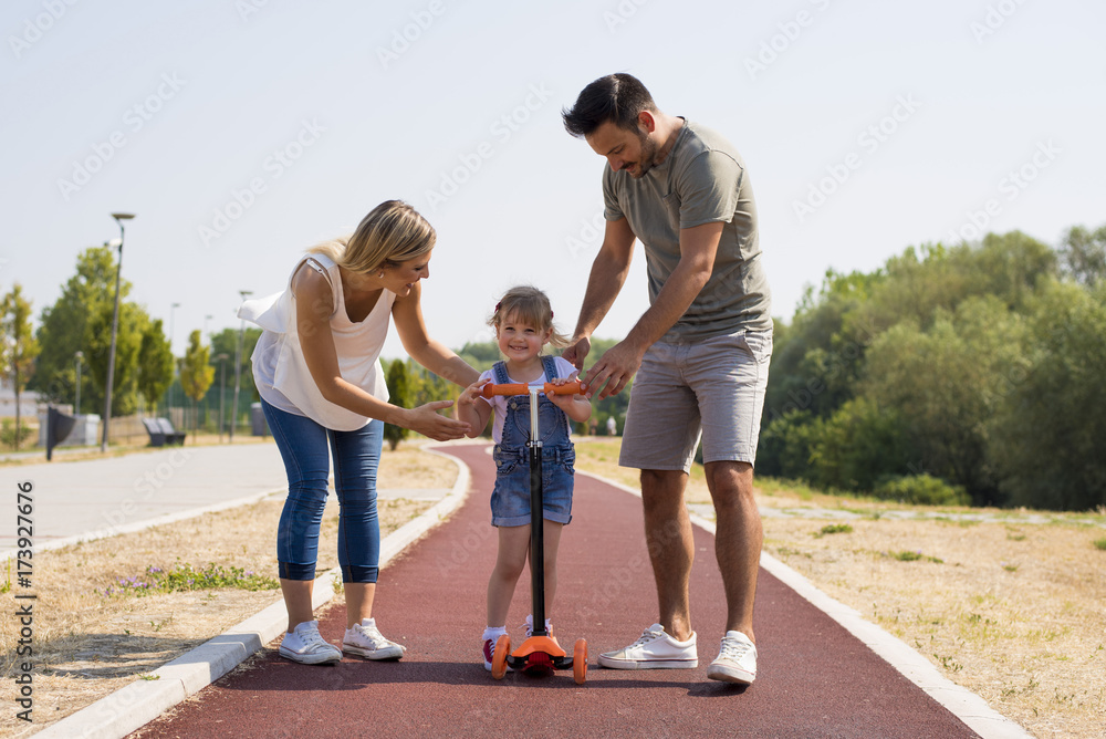Young parents teaching daughter to ride a scooter for the first time Young parents teaching daughter to ride a scooter for the first time