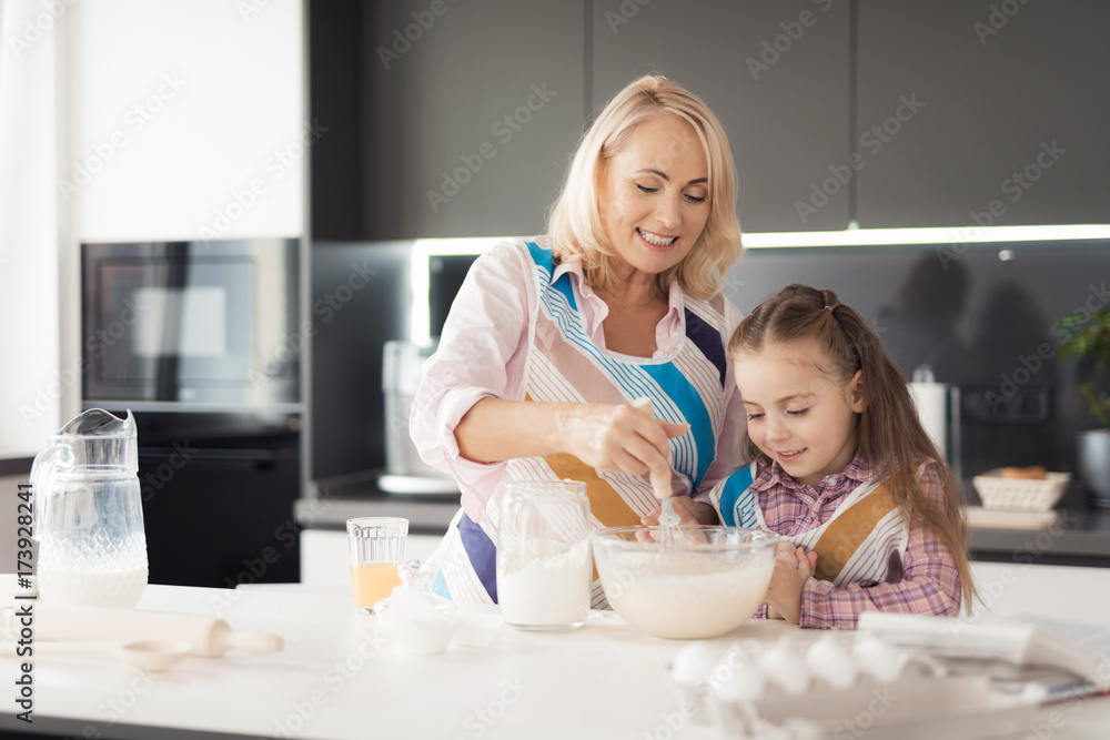 A girl with her grandmother cooks a homemade cake. They put on kitchen aprons and knead the dough