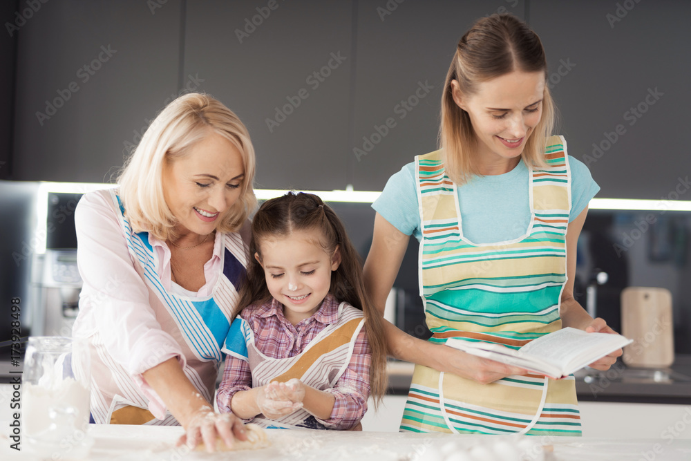 A woman with a recipe book helps a girl and her grandmother prepare a cake A woman with a recipe book helps a girl and her grandmother prepare a cake