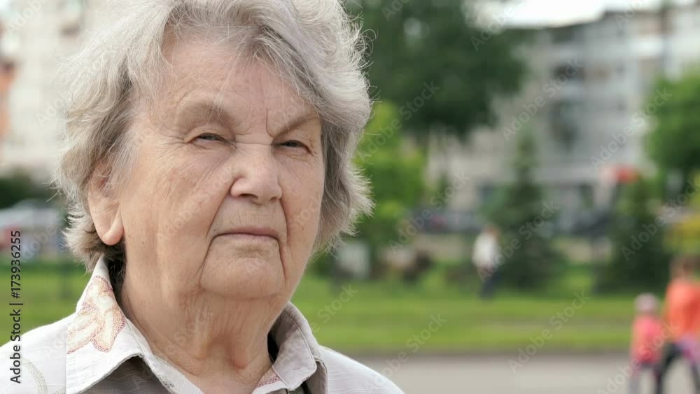 Portrait of serious mature old woman aged 80s outdoors in the background road with passing cars in summer. Slow motion Portrait of serious mature old woman aged 80s outdoors in the background road with passing cars in summer. Slow motion