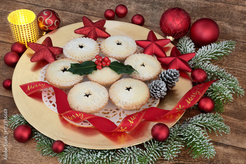 Christmas mince pies with red ribbon on a gold plate with holly, fir, red bauble decorations and candle on oak wood background.