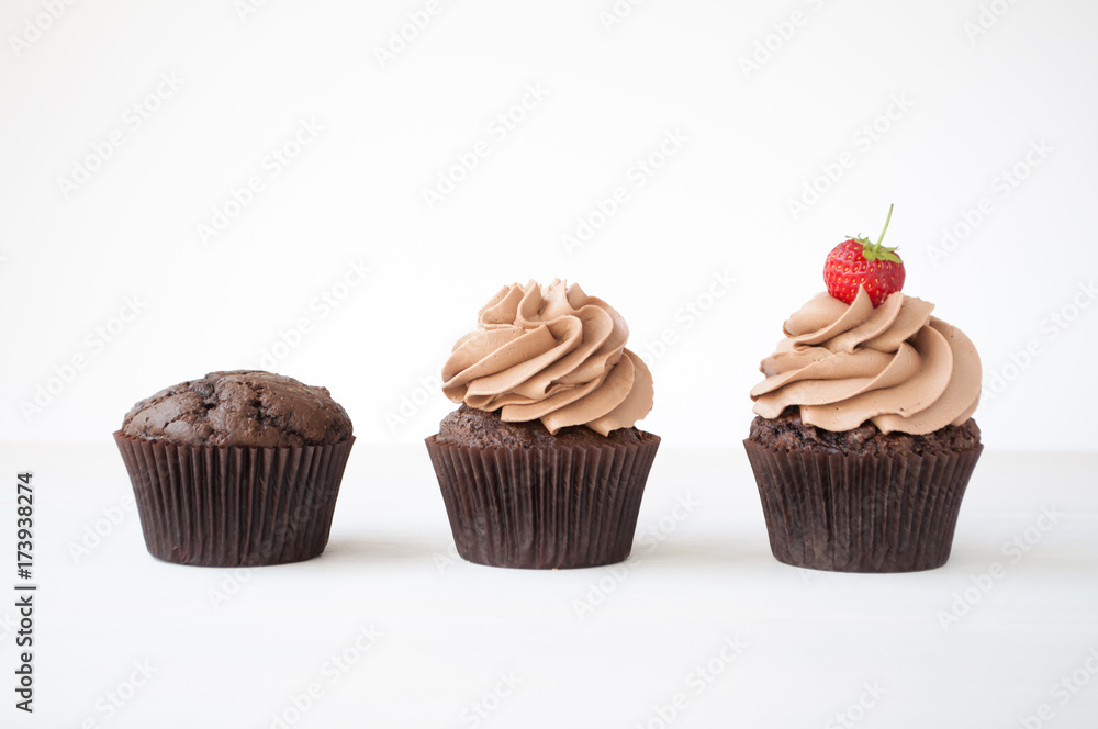 Cupcakes with whipped chocolate cream, decorated fresh strawberry on white wooden table. Picture for a menu or a confectionery catalog, showing different stages of creation.