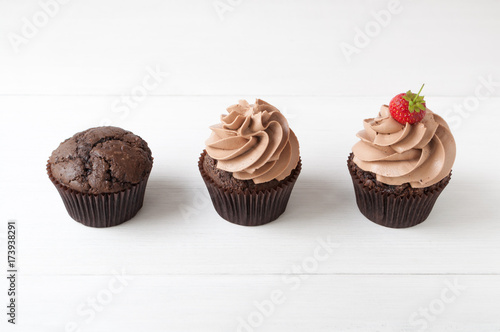 Cupcakes with whipped chocolate cream, decorated fresh strawberry on white wooden table. Picture for a menu or a confectionery catalog, showing different stages of creation.