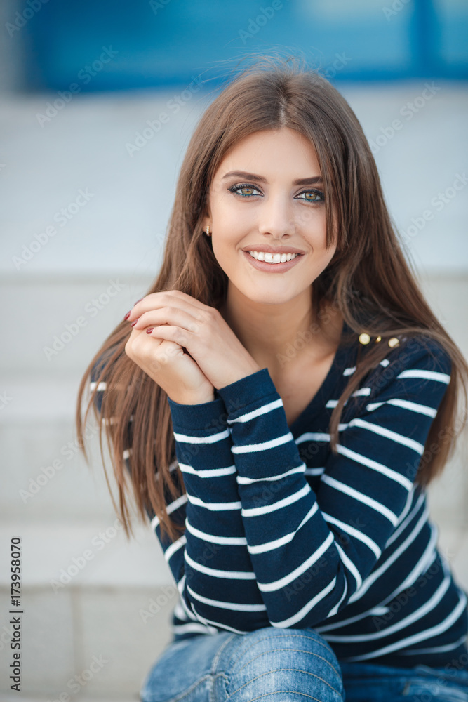 Young beautiful woman brunette with long straight hair and gray-green eyes,dressed in a striped t-shirt and blue jeans,spends time in the city in the spring,sitting on the steps,one in the fresh air